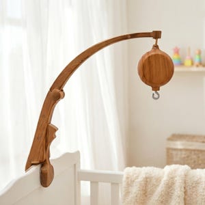 Op de afbeelding: Een houten baby mobile arm met een ronde muziekdoos. De mobiele arm is bevestigd aan een witte wieg. De mobiele arm en muziekdoos zijn gemaakt van lichtbruin hout. De muziekdoos heeft een zilveren haak.