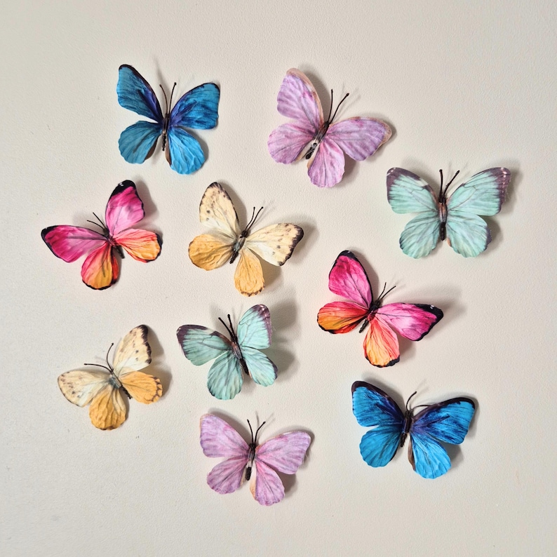 May include: A collection of colorful butterfly decorations in various shades of blue, pink, yellow, and purple. The butterflies have detailed wing patterns and are arranged on a neutral background, creating a whimsical and decorative display.