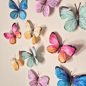 May include: An assortment of colorful butterfly decorations in shades of blue, pink, yellow, and green. The butterflies have detailed wing patterns and are arranged on a neutral background, creating a decorative display.