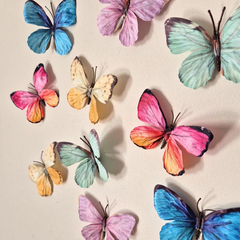 May include: An assortment of colorful butterfly decorations in shades of blue, pink, yellow, and green. The butterflies have detailed wing patterns and are arranged on a neutral background, creating a decorative display.