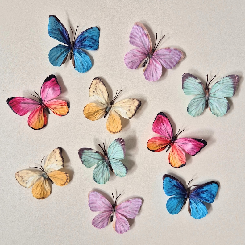 May include: A collection of nine colorful butterfly decorations in various shades of blue, pink, yellow, and purple. The butterflies have detailed wing patterns and are arranged on a neutral background, showcasing their vibrant colors.