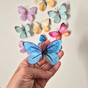 May include: A collection of colorful butterfly decorations. The foreground features a large, vibrant blue butterfly with black accents. Other butterflies in shades of pink, yellow, and blue are arranged in the background, creating a whimsical display.