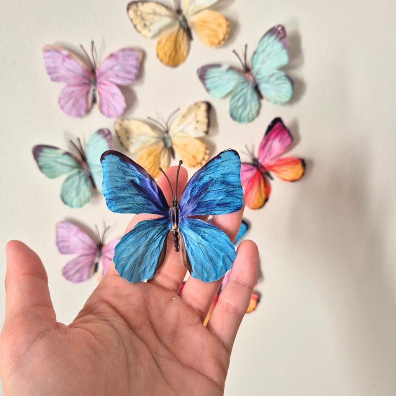May include: A collection of colorful butterfly decorations in various shades of pink, blue, yellow, and orange. One large blue butterfly is held in a hand, with several other butterflies arranged on a white surface. The butterflies have detailed wing patterns.