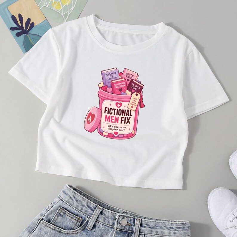 May include: A white short-sleeved t-shirt featuring a graphic of a pink pill bottle filled with small books. The bottle is labeled FICTIONAL MEN FIX and includes the text take one more chapter daily, with small hearts and a tag reading just one more chapter.