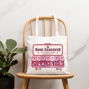May include: A white tote bag featuring a graphic of a pink pill organizer with seven compartments labeled Monday through Sunday. The text reads BOOK HANGOVER, emotionally destroyed by fictional men, with small heart icons and pink capsules.