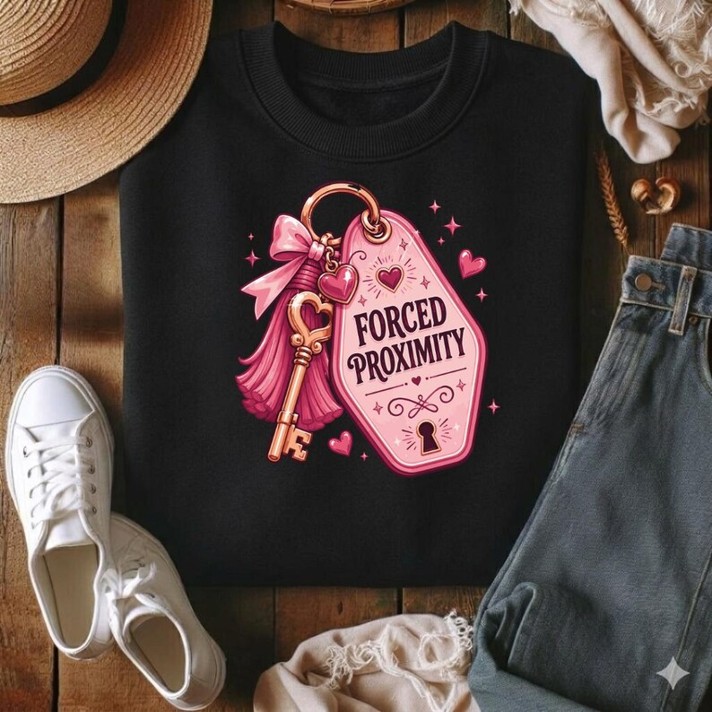 May include: A black crewneck sweatshirt featuring a graphic print of a pink motel-style keychain tag with the text FORCED PROXIMITY, a gold-colored key, a pink bow, and small heart accents. The design is centered on the front of the garment.