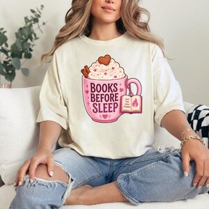 May include: A cream-colored t-shirt featuring a graphic of a pink mug filled with whipped cream, cinnamon sticks, and a heart-shaped cookie. The mug displays the text "BOOKS BEFORE SLEEP" and a small book charm with the words "just one more chapter".
