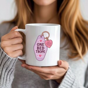 May include: A white ceramic mug featuring a graphic of a light purple motel-style keychain with the text ONE BED TROPE and small pink hearts. Attached to the keychain is a smaller pink heart charm with the text book boyfriend energy.