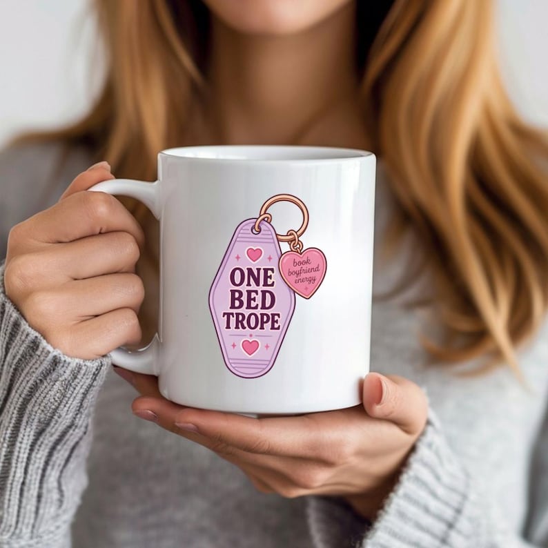 May include: A white ceramic mug featuring a graphic of a light purple motel-style keychain with the text ONE BED TROPE and small pink hearts. Attached to the keychain is a smaller pink heart charm with the text book boyfriend energy.