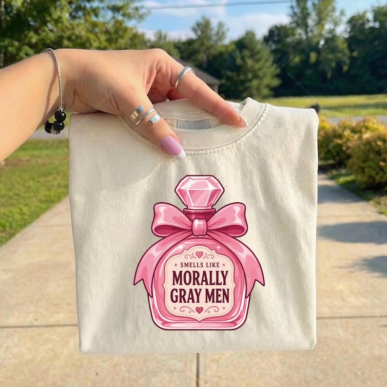 May include: A cream-colored t-shirt featuring a graphic of a pink perfume bottle with a large bow and a diamond-shaped cap. The text on the bottle reads, "SMELLS LIKE MORALLY GRAY MEN," surrounded by small decorative heart and star accents.