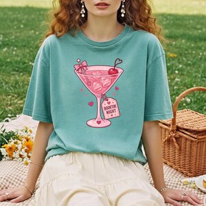 May include: A sage green short-sleeve t-shirt featuring a graphic of a pink martini glass filled with heart-shaped ice cubes, a cherry, and a pink bow. A pink tag attached to the glass stem displays the text "BOOKTOK NIGHT" in bold letters.