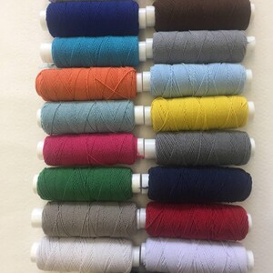 以下が含まれることがあります： 白のプラスチックコアに巻かれた、カラフルな糸巻きのコレクション。糸は青、オレンジ、緑、グレーなど様々な色合いです。糸巻きは整然と並べられており、色の多様性を示しています。