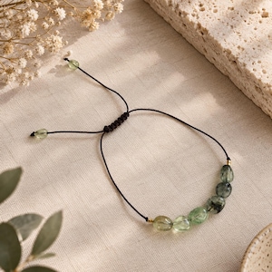Puede incluir: Una pulsera con un cordón negro fino y un cierre de nudo corredizo ajustable. El diseño incluye una serie de cuentas de piedra pulidas, lisas y translúcidas en tonos verde y verde oscuro, acentuadas por pequeñas cuentas de metal dorado.