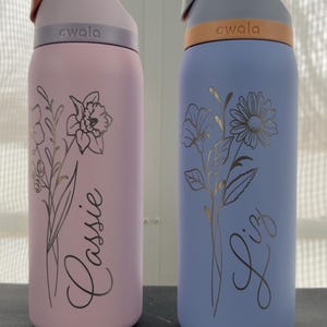May include: Two insulated water bottles, one pink and one blue, each with a floral design and a name. The pink bottle says "Lassie" and the blue bottle says "Liz". Both bottles have a gray lid and a carrying handle.
