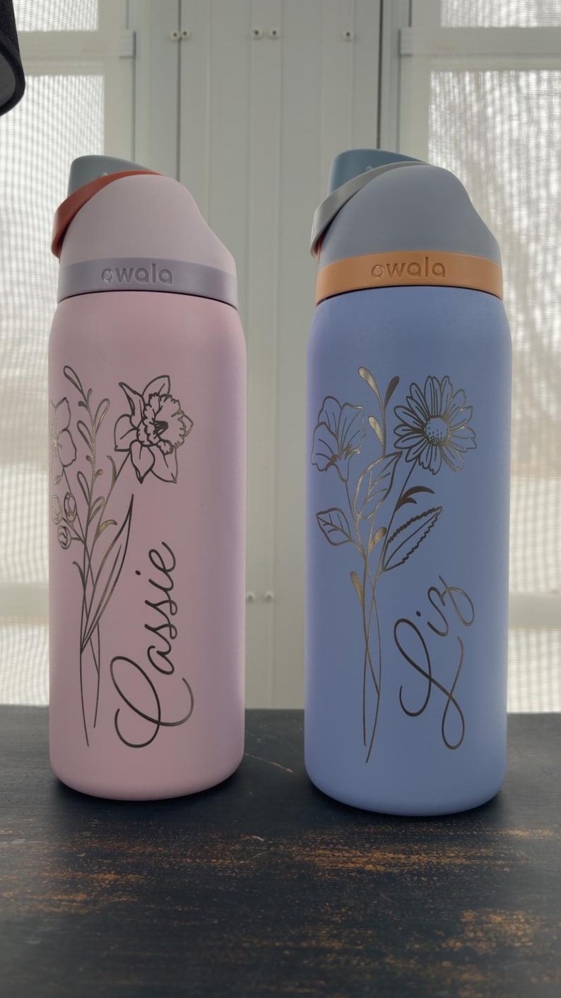 May include: Two insulated water bottles, one pink and one blue, each with a floral design and a name. The pink bottle says "Lassie" and the blue bottle says "Liz". Both bottles have a gray lid and a carrying handle.