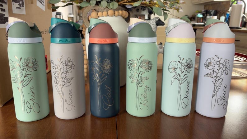 May include: Six colorful water bottles with floral designs and names. Each bottle has a different color lid and base, with names like "Erin" and "Staci" in script. The bottles are on a wooden surface.