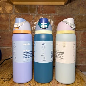 May include: Three Owala insulated water bottles in pastel colors: lavender, teal, and mint green. Each bottle has a FreeSip spout and a built-in straw. The bottles have a capacity of 32 oz (946 ml). The text on the bottles says, "Hi there. I'm leak proof. I'm insulated. And I have a built-in straw."