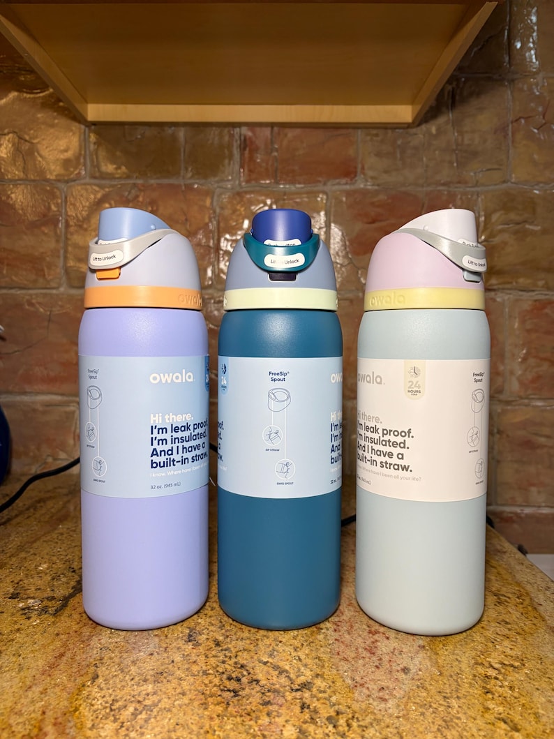 May include: Three Owala insulated water bottles in pastel colors: lavender, teal, and mint green. Each bottle has a FreeSip spout and a built-in straw. The bottles have a capacity of 32 oz (946 ml). The text on the bottles says, "Hi there. I'm leak proof. I'm insulated. And I have a built-in straw."
