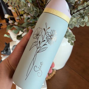 May include: A light blue Owala water bottle with a floral design and the name "Sue" in cursive. The bottle has a light pink lid, a yellow band, and a silver floral accent. The bottle is approximately 10 inches tall.