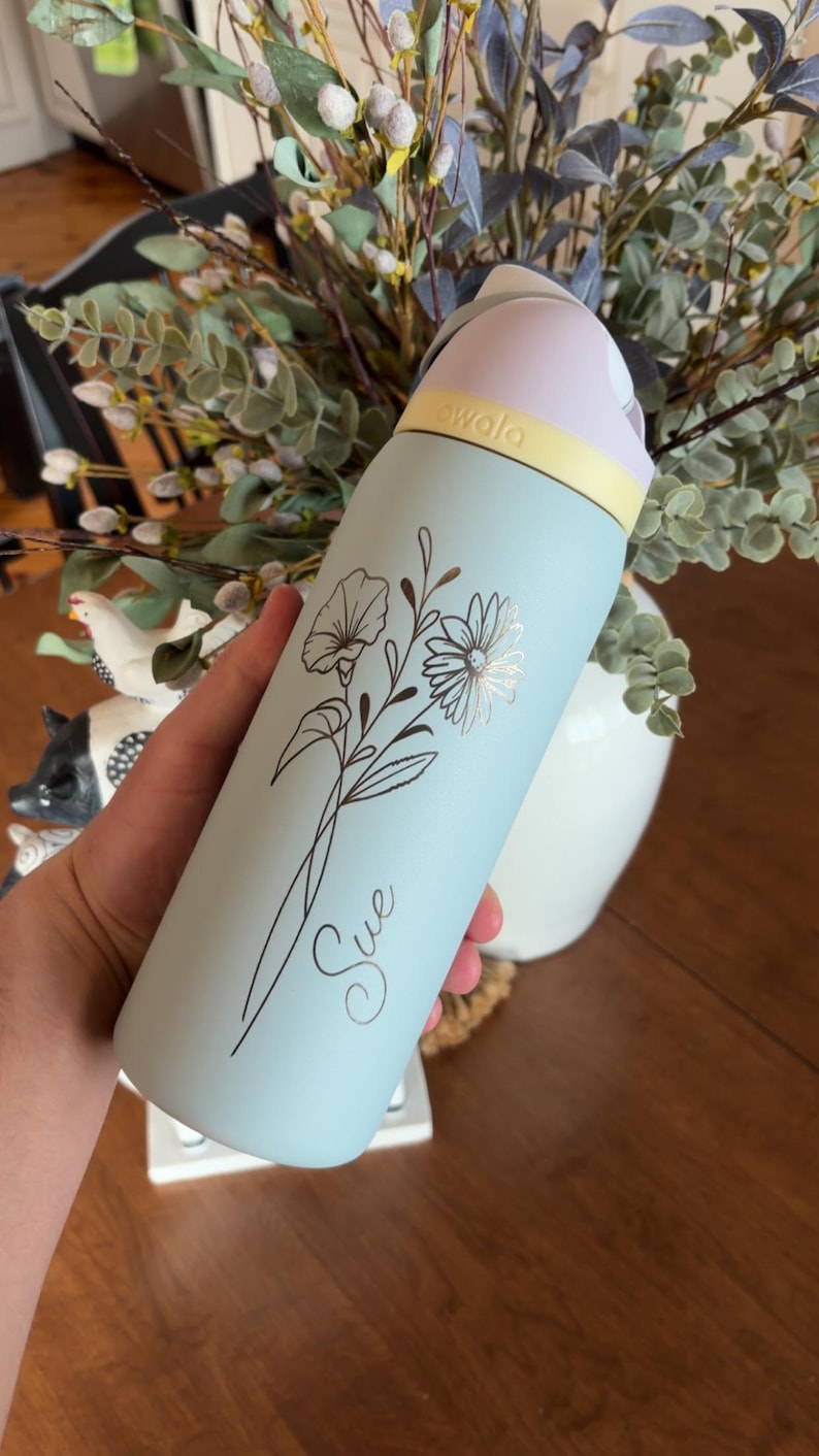 May include: A light blue Owala water bottle with a floral design and the name "Sue" in cursive. The bottle has a light pink lid, a yellow band, and a silver floral accent. The bottle is approximately 10 inches tall.