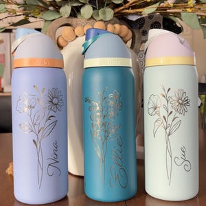 May include: Three insulated water bottles in pastel colors: lavender, teal, and mint green. Each bottle features a floral design and a name in cursive script. The bottles have colored lids and bands, with the brand name "owala" visible.