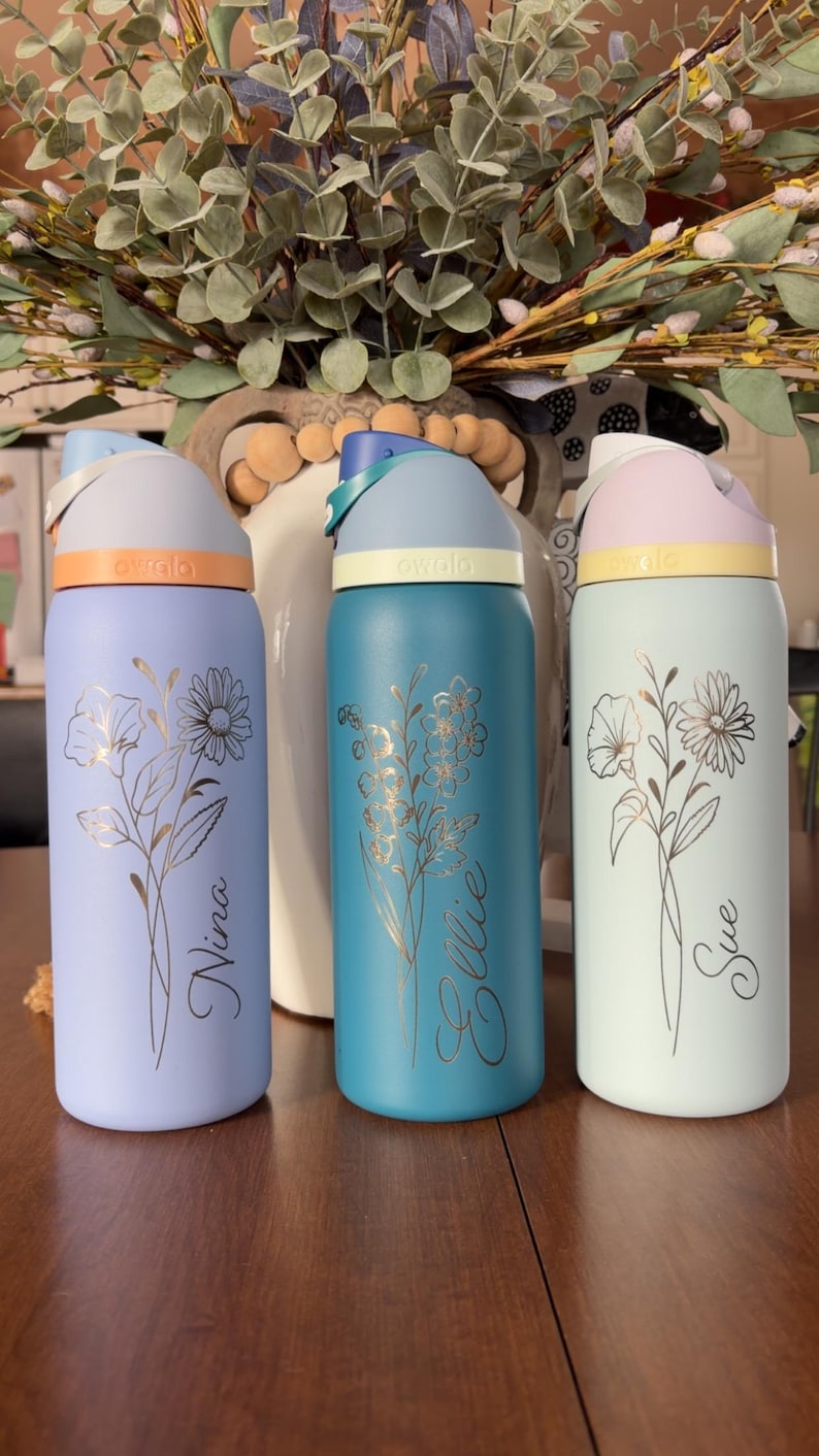 May include: Three insulated water bottles in pastel colors: lavender, teal, and mint green. Each bottle features a floral design and a name in cursive script. The bottles have colored lids and bands, with the brand name "owala" visible.
