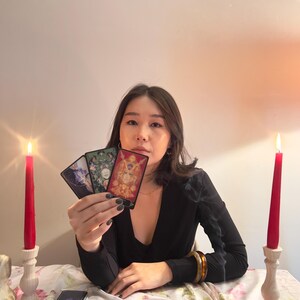 May include: Three rectangular tarot cards held in a hand, featuring intricate illustrations of figures and nature. Two red taper candles in white holders stand on either side of a floral tablecloth covered with a deck of cards and incense sticks.