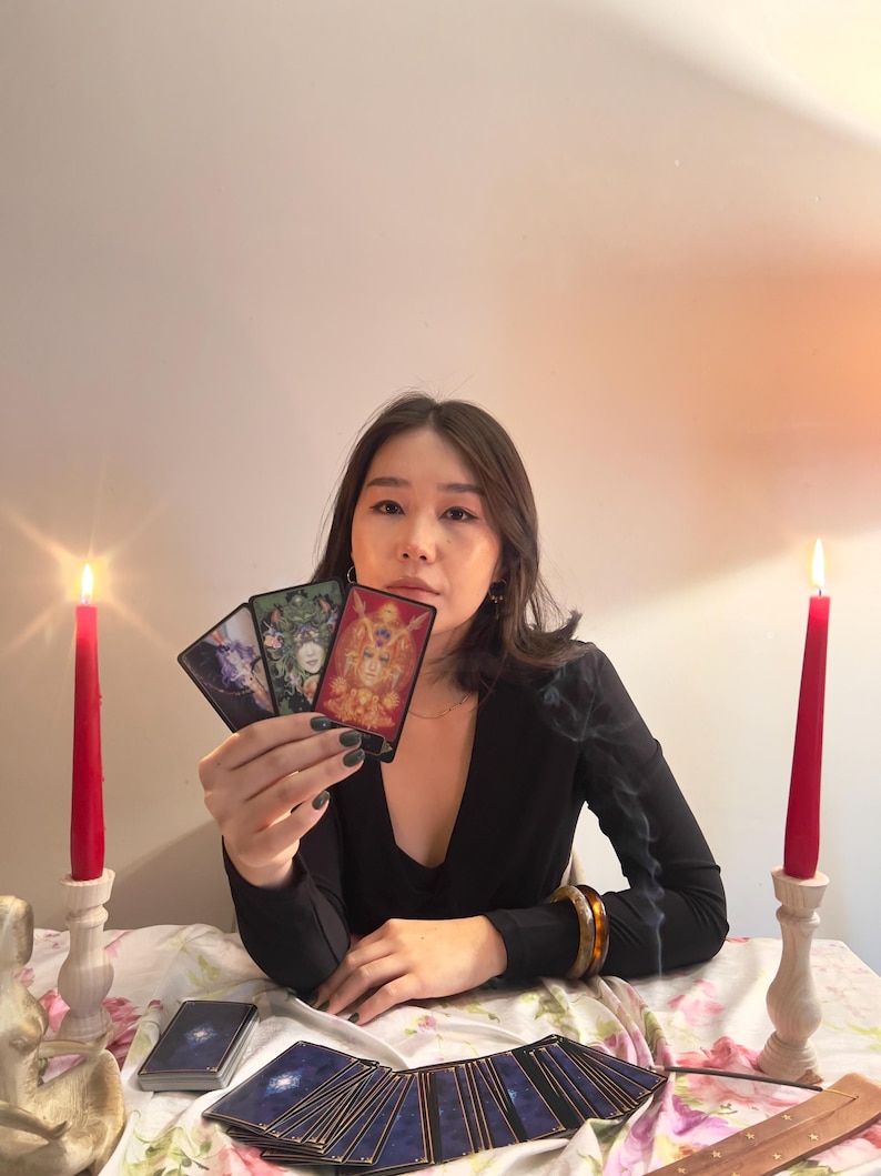 May include: Three rectangular tarot cards held in a hand, featuring intricate illustrations of figures and nature. Two red taper candles in white holders stand on either side of a floral tablecloth covered with a deck of cards and incense sticks.