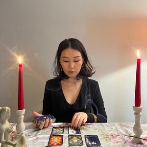 May include: A set of tarot cards arranged in a spread on a floral tablecloth, flanked by two red taper candles in wooden holders. A small figurine and an incense holder are present, with a deck of cards held in one hand and another being placed.