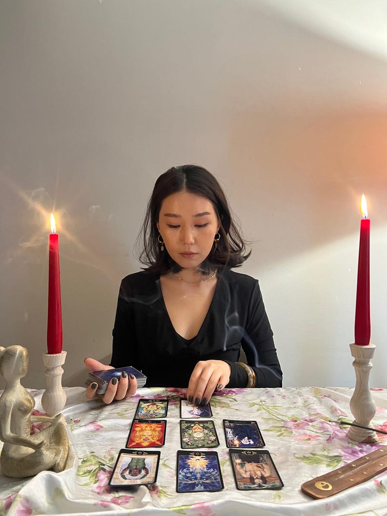 May include: A set of tarot cards arranged in a spread on a floral tablecloth, flanked by two red taper candles in wooden holders. A small figurine and an incense holder are present, with a deck of cards held in one hand and another being placed.
