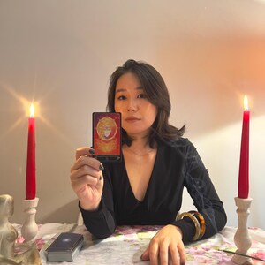 May include: A rectangular tarot card with a red and gold design featuring a central figure, held upright. The card displays the text "THE EMPEROR" at the bottom. The scene includes two red candles in wooden holders and three additional tarot cards laid out.