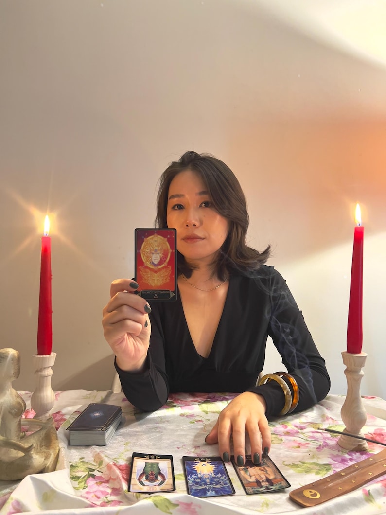 May include: A rectangular tarot card with a red and gold design featuring a central figure, held upright. The card displays the text "THE EMPEROR" at the bottom. The scene includes two red candles in wooden holders and three additional tarot cards laid out.
