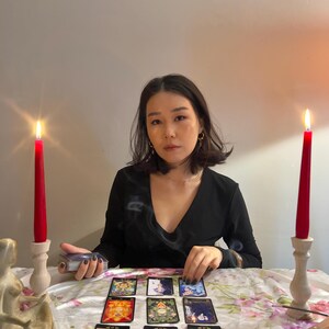 May include: A set of nine tarot cards arranged in a grid on a white floral tablecloth, flanked by two tall red taper candles in light-coloured wooden holders. A small incense burner sits to the right, and a deck of cards is held in the left hand.