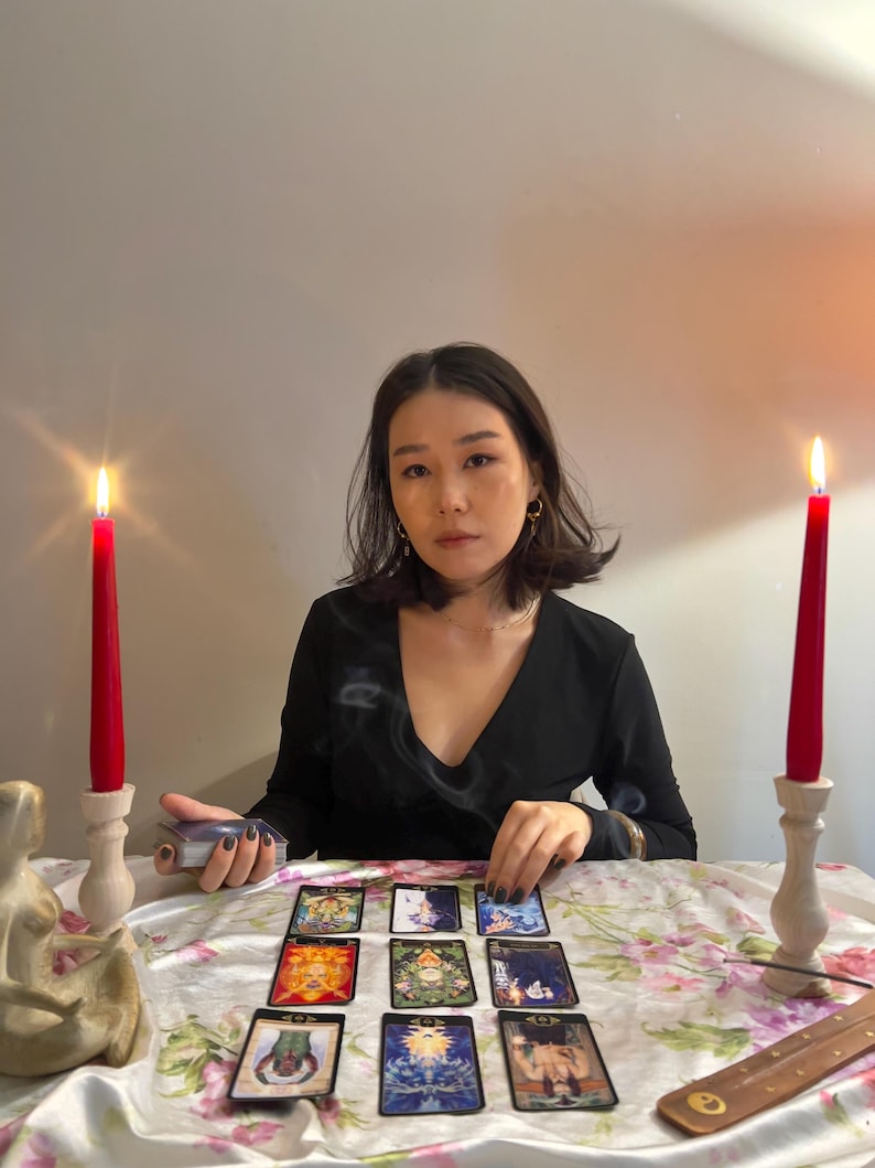 May include: A set of nine tarot cards arranged in a grid on a white floral tablecloth, flanked by two tall red taper candles in light-coloured wooden holders. A small incense burner sits to the right, and a deck of cards is held in the left hand.