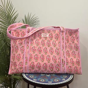 May include: A rectangular quilted tote bag in a soft pink hue featuring a repeating floral pattern in darker pink and green. The bag has white and pink striped fabric handles and trim, with a small white label reading sweetnest studio.