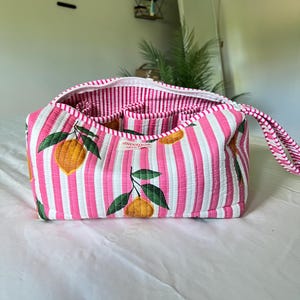 May include: A quilted fabric pouch featuring a pattern of yellow lemons with green leaves on a background of vertical pink and white stripes. The bag has a zippered opening, a side handle, and a small white label reading Sweetnest Studio.