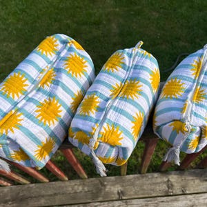 Peut inclure: Trois pochettes en tissu matelassé de tailles différentes, présentant un fond blanc avec des rayures horizontales bleu clair et un motif répétitif de soleils jaune vif. Chaque pochette possède une fermeture éclair blanche avec un pompon.