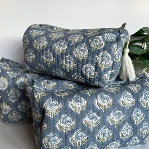 May include: A set of three rectangular quilted pouches in a dusty blue fabric featuring a repeating white and light green floral block print pattern. Each pouch has a zip closure with a white tassel pull attached to the metal zip slider.