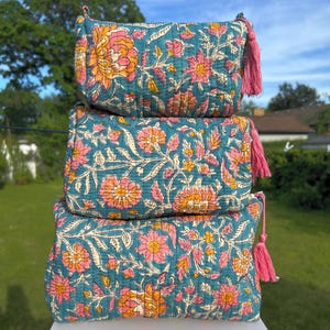 Peut inclure: Une pile de trois trousses rectangulaires matelassées en tissu bleu sarcelle avec un motif floral répétitif dans des tons rose, orange et crème. Chaque trousse possède un pompon rose assorti attaché à la tirette de la fermeture éclair.