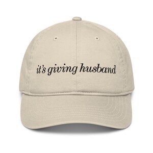 May include: A beige baseball cap with a curved brim. The cap features the phrase "it's giving husband" in black script across the front. The hat is made of a soft material and has a classic, casual style.