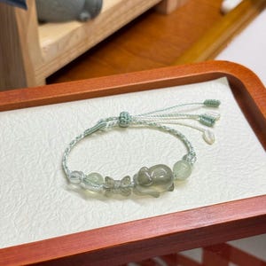 May include: A light green bracelet with a sliding knot closure. The bracelet features a series of light green beads and charms, including a small pig and a butterfly. The bracelet is displayed on a white surface within a wooden tray.