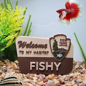 May include: A decorative sign for a fish tank with the text "Welcome to my habitat" and "Fishy". A red and white Betta fish swims near the sign. The sign also has a Fish Tank Service emblem.