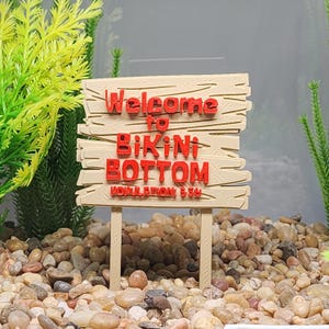 May include: A decorative sign with the words "Welcome to Bikini Bottom" in red lettering. The sign is made of beige-colored wood-like material and is mounted on two beige posts. The sign is placed in a bed of small stones.