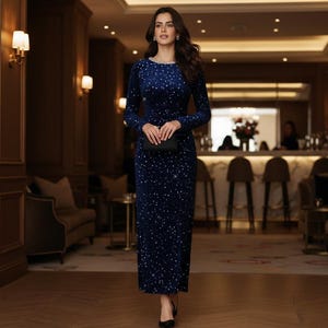 May include: A long, navy blue, floor-length dress with long sleeves and a round neckline. The dress is embellished with small, scattered, silver-colored sequins. A black clutch is held in front of the dress. The setting is a well-lit interior.