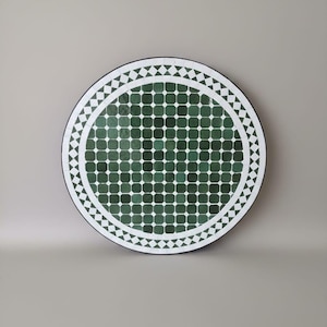 May include: Round decorative table top with a mosaic tile design. The center features a grid of dark green squares. A white border with diamond shapes surrounds the green tiles. The outer edge is black.