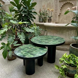 May include: Two round side tables with black cylindrical bases and tops covered in a mosaic of small, glossy green tiles. The tables are of different heights, with the larger one positioned behind the smaller one, both featuring a smooth finish.