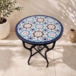 Può includere: Un tavolino rotondo con piano in mosaico caratterizzato da un motivo geometrico a stella nei toni del blu, marrone e bianco. Il tavolo ha una struttura in metallo nero con quattro gambe curve collegate da un supporto centrale decorativo.