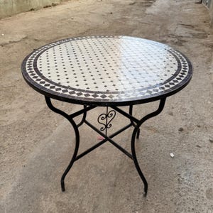 May include: A round table featuring a mosaic tile top with a white and black geometric pattern. The table is supported by a black metal frame with four curved legs and a central decorative scrollwork element connecting the base supports.