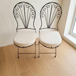 May include: Two wrought iron chairs with cream-colored cushions. The backs feature an ornate, scrolled design. The chairs are set on a light-colored wood floor, with a neutral wall in the background. The chairs are a classic design.