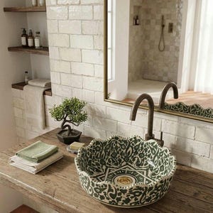 Puede incluir: Un lavabo de cerámica redondo con borde festoneado, decorado con un patrón floral en verde oscuro y blanco. El lavabo cuenta con un desagüe dorado y un grifo de cuello de cisne en bronce oscuro, sobre una encimera de madera rústica.